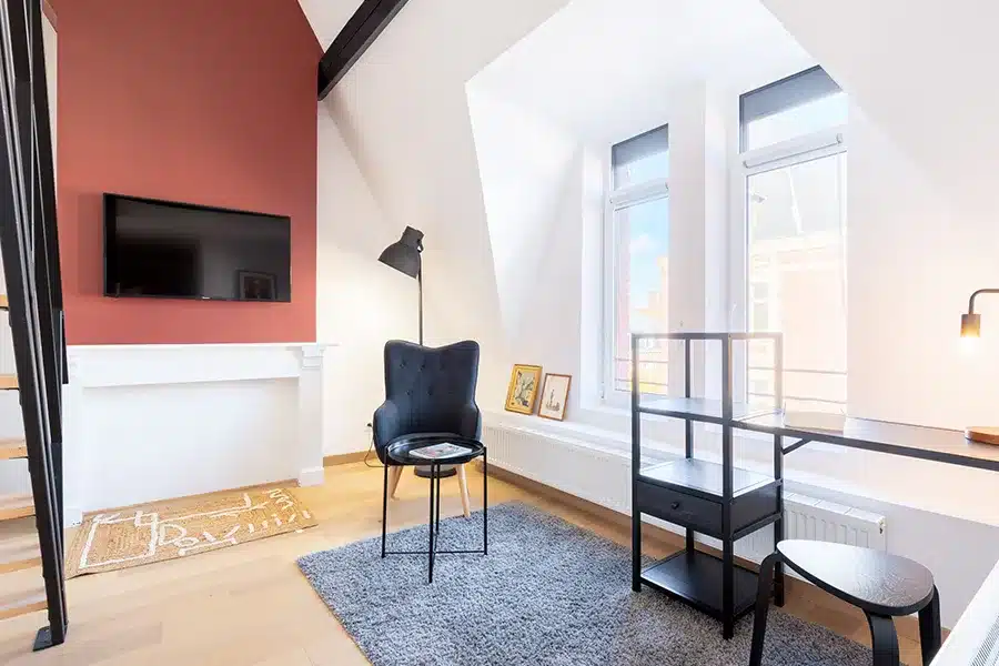 Salon mansardé avec mur rouge brique, télévision et fauteuil bleu nuit — Maison Barbieux, coliving IvyNest à Roubaix