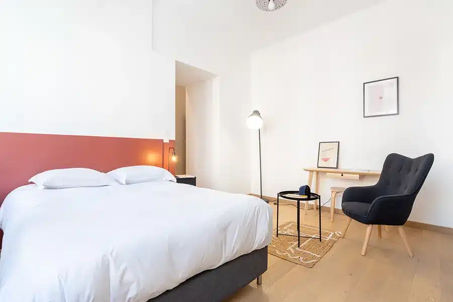 Chambre spacieuse avec lit double, tête de lit rouge brique et fauteuil gris — Maison Barbieux, coliving IvyNest à Roubaix