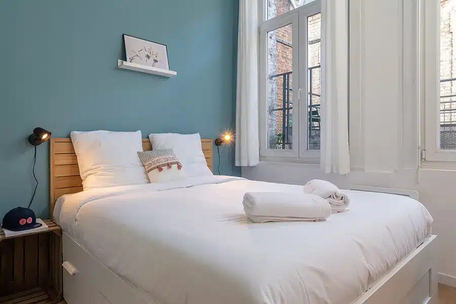 Chambre avec lit double, mur bleu canard et fenêtre haute à rideaux blancs — Maison Colbert, coliving IvyNest à Lille